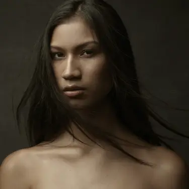 Boundary-pushing model headshot with dark hair and olive toned skin, bare shoulders, serious gaze against dark neutral background, headshot photographer 312 Elements Headshot Photography | Chicago, IL