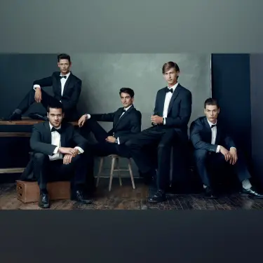 Five men in formal black tuxedos posed together against blue-gray studio backdrop, arranged at varying heights creating dynamic group composition, image by Chicago, IL portrait photographer 312 Elements Headshot Photography