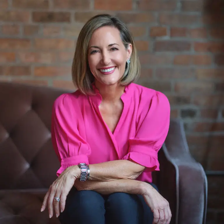 Susan Guthrie in vibrant fuchsia blouse seated on gray chair against soft brick wall backdrop, professional headshot in Chicago, photographed by Chicago, IL headshot photographer 312 Elements Headshot Photography
