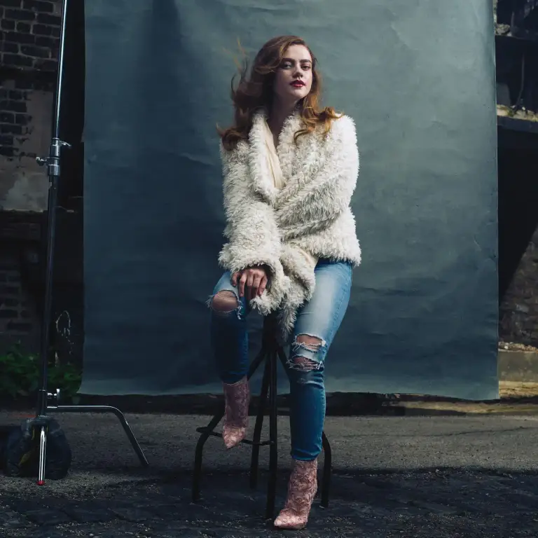 Actress Bella Ortiz portrait in Chicago alley wearing cream sweater and glasses, seated on black chair against blue-gray wall, photographed by Chicago, IL portrait photographer 312 Elements Headshot Photography