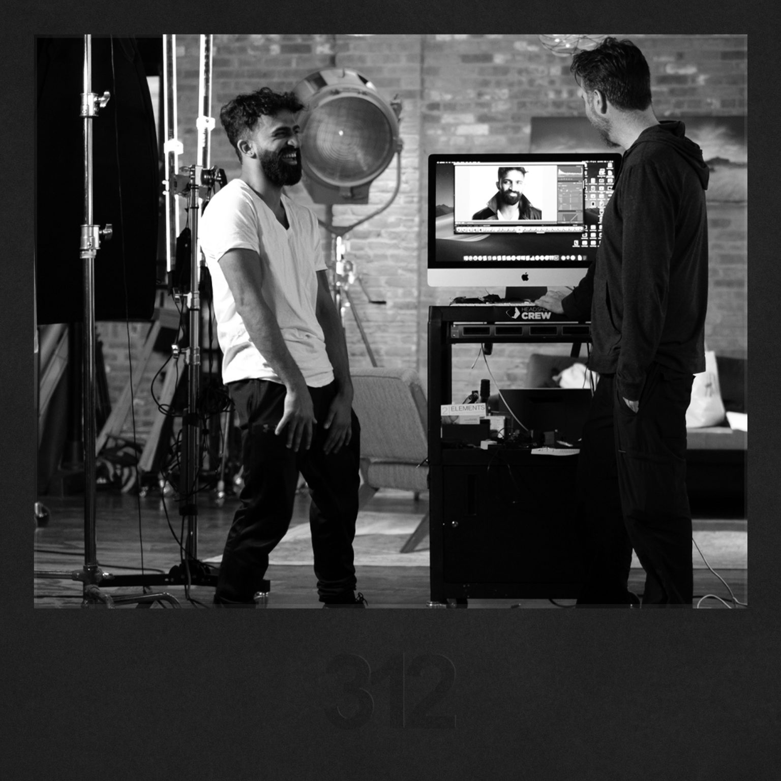 A cheerful client shares a laugh with their photographer next to a computer displaying headshot images, engaging in a conversation about rescheduling.