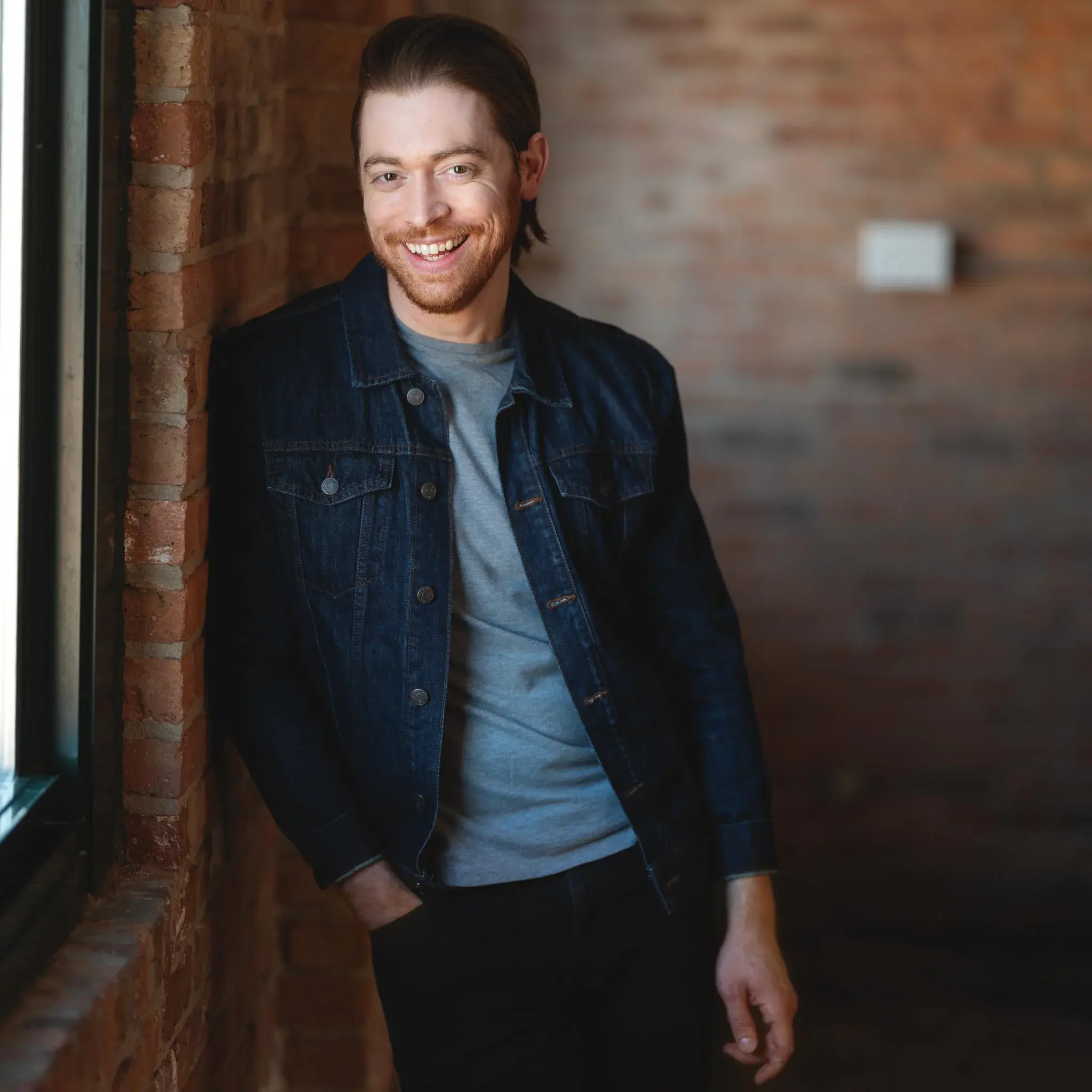 High-quality actor headshot of a smiling individual positioned against a rustic brick wall, ideal for casting calls and promotional materials.